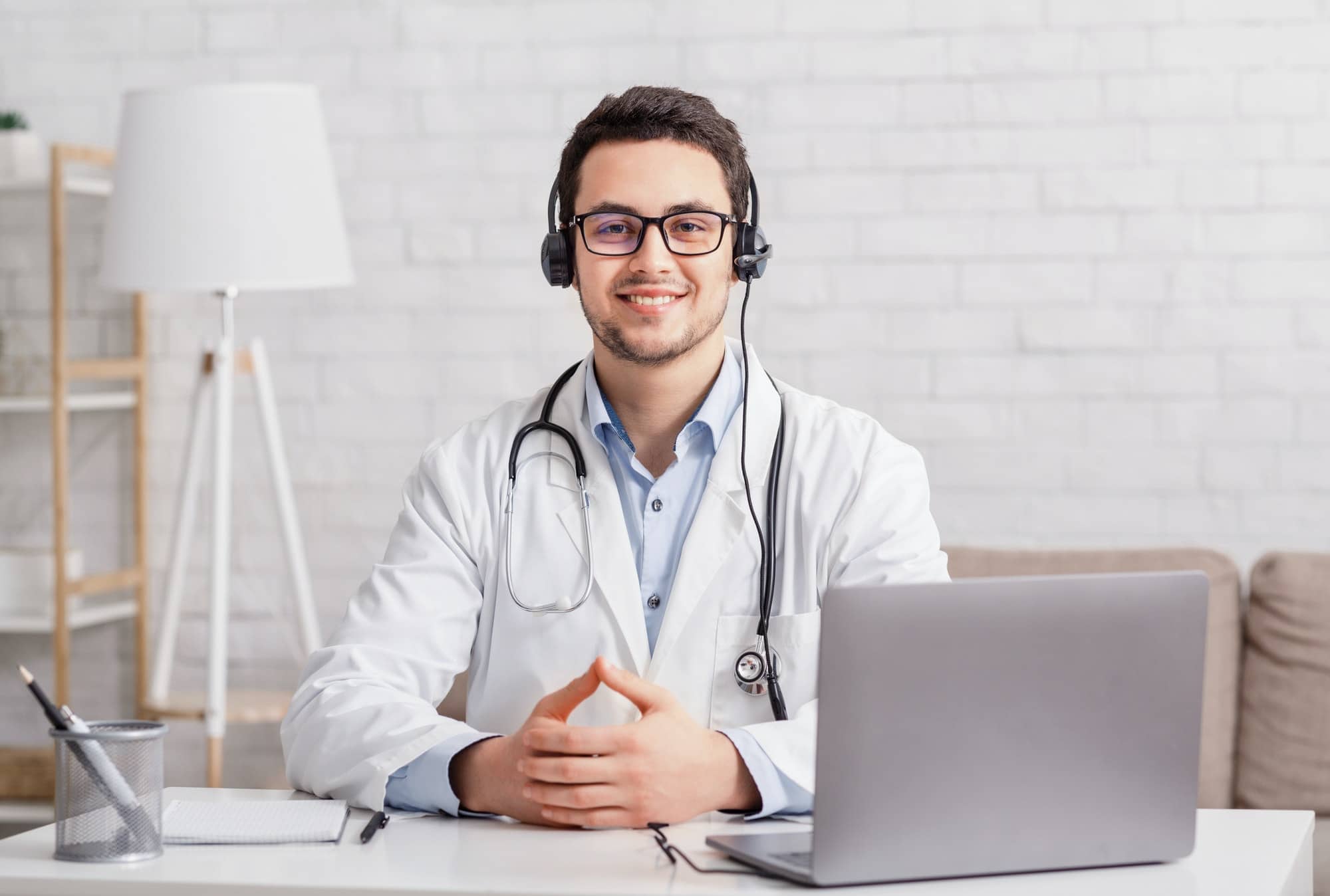 Health blog. Young doctor in headphones sitting at table with laptop