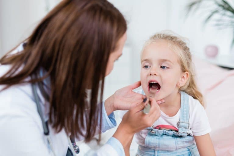 Sick child opening mouth while looking at doctor examining her sore throat